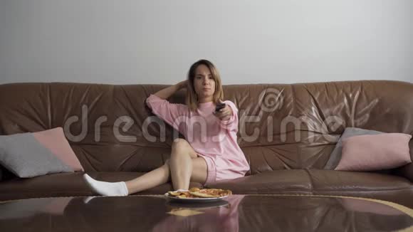 无聊的年轻女人坐在棕色的皮革沙发上看电视家庭主妇用遥控器切换频道视频的预览图