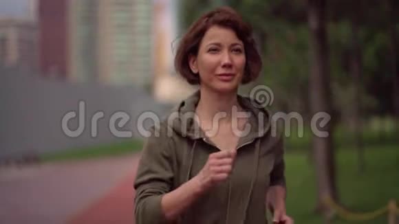 慢动作吊环跟踪年轻有魅力的穿着帽衫的女人在绿色城市公园跑步视频的预览图