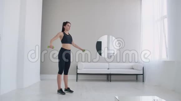 一个女人用哑铃举起手臂在她那间浅色的白色公寓里靠在沙发和窗户上做视频的预览图