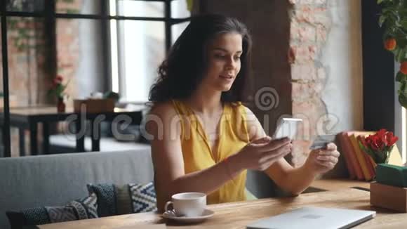 兴奋的女孩独自坐在咖啡馆里用智能手机和信用卡在线支付视频的预览图