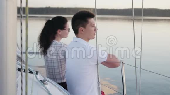 年轻的已婚夫妇穿着红裤子坐在一艘白色的帆船旁边在黑暗平静的水面上航行视频的预览图