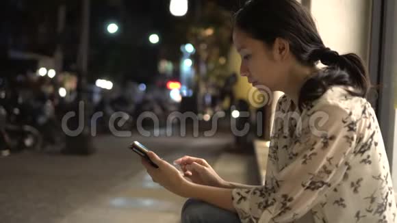 一个亚洲女人坐在城市街边的一晚使用智能手机视频的预览图