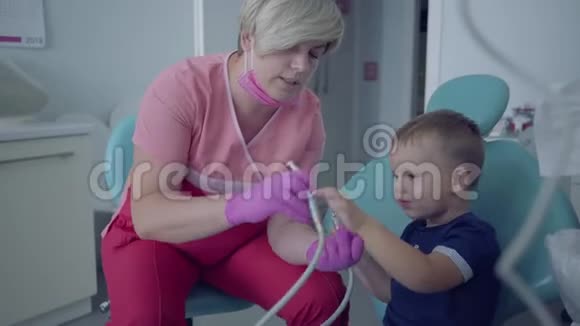 牙医戴着医用口罩和手套准备检查坐在椅子上的无忧无虑的小男孩的牙齿女性视频的预览图