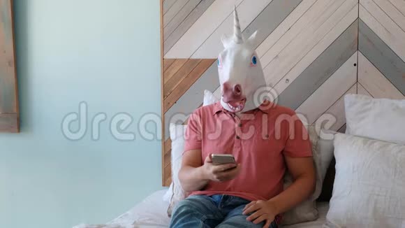 奇怪的搞笑视频戴着独角兽面具的男人躺在床上看着智能手机视频的预览图