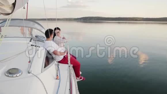 年轻的已婚夫妇穿着红裤子坐在一艘白色的帆船旁边在黑暗平静的水面上航行视频的预览图