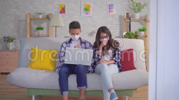 厌倦了一对寒冷的年轻夫妇他们坐在沙发上戴着保护性医疗面具用笔记本电脑视频的预览图