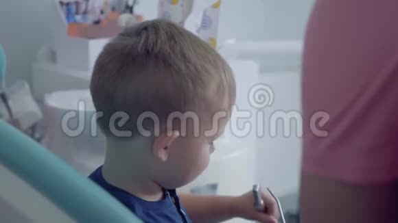 爱的牙医用双手检查坐在椅子上的无忧无虑的小男孩的牙齿给他看一张镜子特写视频的预览图