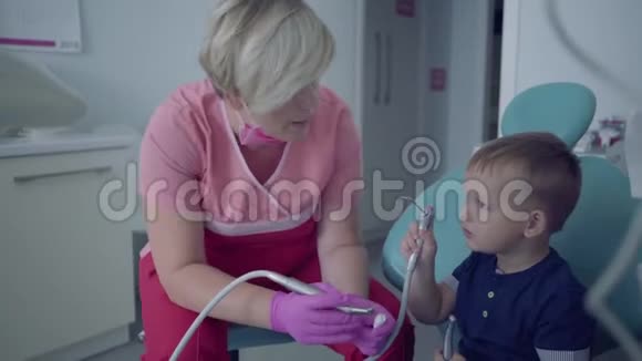 牙医戴着医用口罩和手套准备检查坐在椅子上的无忧无虑的小男孩的牙齿女性视频的预览图