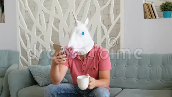 奇怪的搞笑视频戴着独角兽面具的男人坐在沙发上拿着智能手机喝咖啡视频的预览图