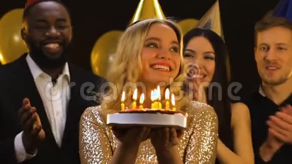 戴着生日帽的金发女人微笑着在蛋糕上吹蜡烛朋友们鼓掌视频的预览图