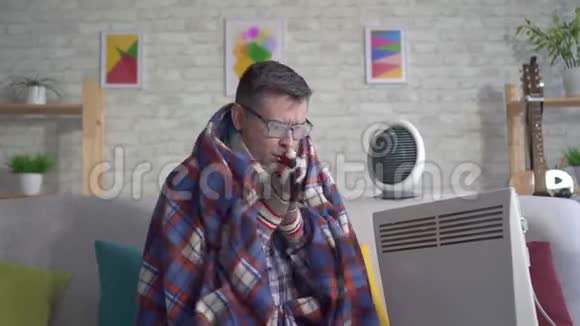 裹在毯子里的冷冻中年男子被加热到电加热器旁边看着摄像机视频的预览图