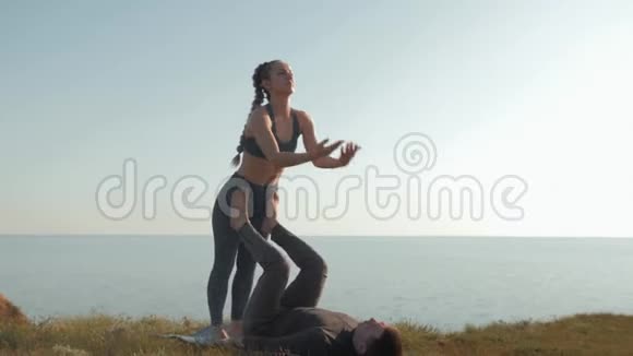 户外练习瑜伽身穿运动服的女孩正在草地上平衡她的男伴的腿和手视频的预览图