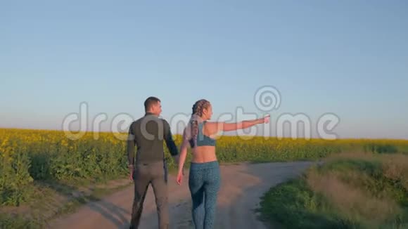 运动休闲年轻夫妇带着卡尼卡隆辫子和运动身体在盛开的油菜花田里散步视频的预览图