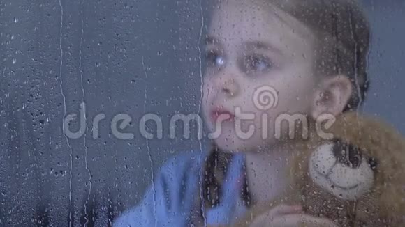 孤独可爱的女孩在雨窗后的孤儿院拥抱泰迪熊做梦视频的预览图