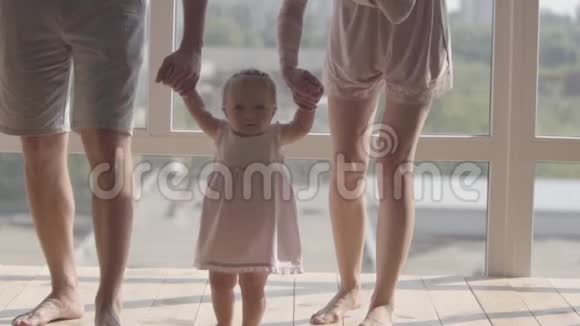 无法辨认的男人和女人在家牵着可爱的婴儿的手在孩子的帮助下迈出第一步视频的预览图