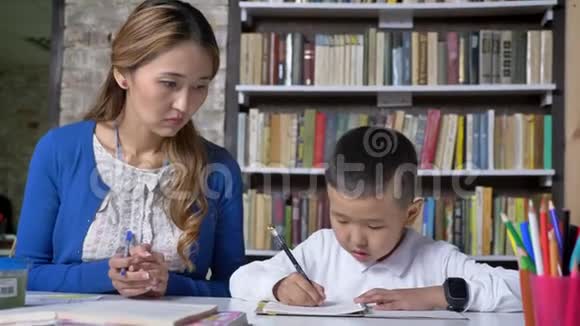 年轻的母亲帮助儿子学习女人和孩子说话坐在桌子后面亚洲孩子做作业看书视频的预览图