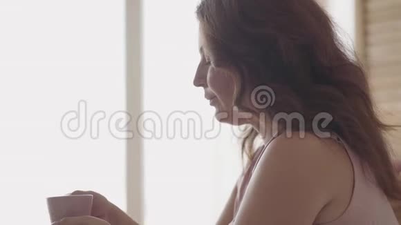 肖像可爱的成熟女人早上坐在床上喝茶或咖啡微笑的女士在家放松太阳视频的预览图