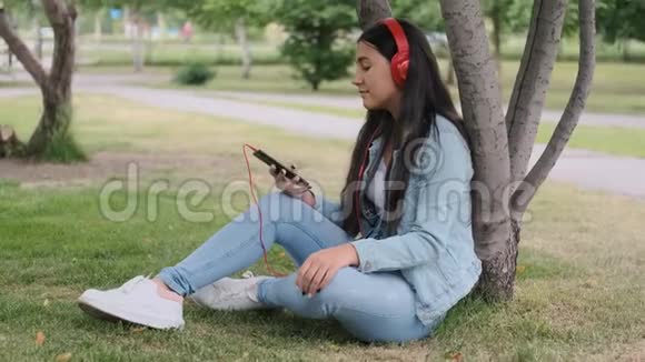 女孩坐在公园的一棵树旁听着耳机里的音乐视频的预览图