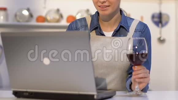 穿着围裙的女士喝着酒在笔记本电脑上选择网上食谱做晚餐视频的预览图
