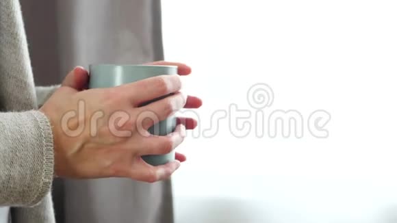女孩手里拿着一杯热茶里面有蒸汽一个女人站在窗口喝热饮料视频的预览图