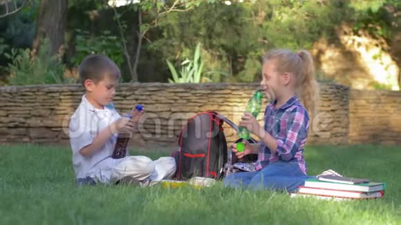 快乐的年轻朋友一起在校园午餐时从坐在草坪上背包旁的瓶子里喝果汁视频的预览图