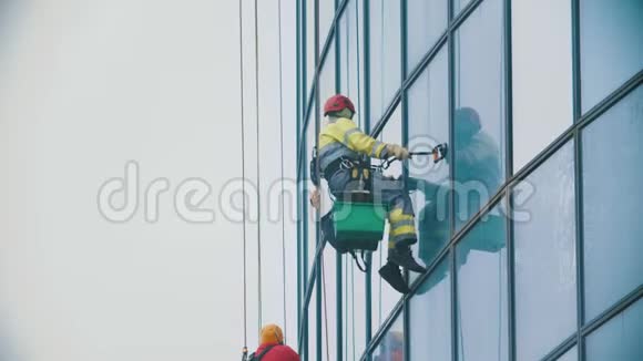 一名穿着黄色工作服的工人挂在绳子上擦拭一座商业摩天大楼的外墙视频的预览图