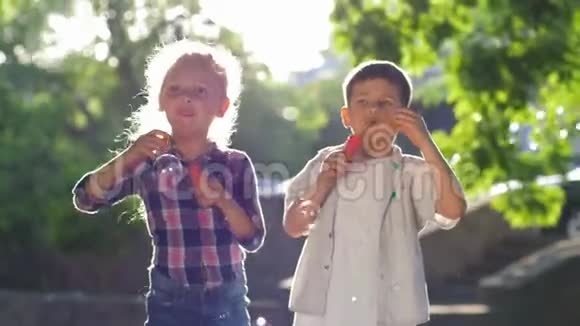 快乐的童年女孩和男孩在公园里吹肥皂泡在背光下的新鲜空气中玩耍视频的预览图