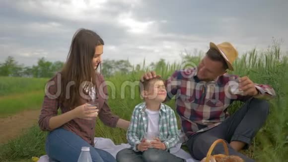 快乐的童年微笑的妈妈和爸爸和小儿子在家庭野餐时喝牛奶在绿野里拥抱户外视频的预览图
