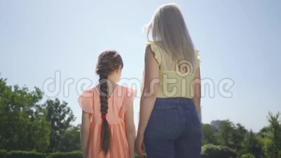 从后面可以看到金发母亲和她的小女儿一起在夏天的花园或公园里度过的时光拥抱的女孩视频的预览图