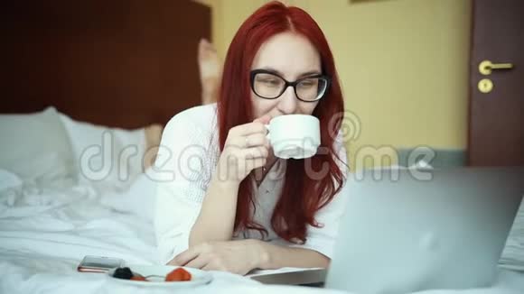 一个穿着浴袍躺在床上喝着茶的生姜女人用笔记本电脑工作自由职业视频的预览图