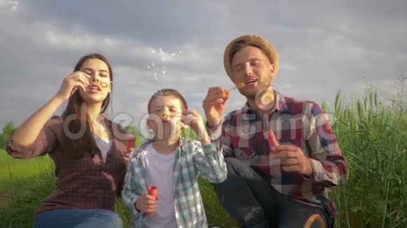 家庭的乐趣快乐的爸爸妈妈和儿子一起吹泡泡在绿色的田野上休息时微笑视频的预览图