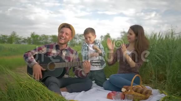 快乐的爸爸演奏乐器而妈妈和儿子一起在大自然的家庭野餐中尽情享受和鼓掌视频的预览图