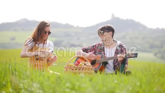 一对年轻夫妇坐在一片明亮的绿色草地上一个男人在弹吉他而他的女朋友在杯子里倒酒视频的预览图