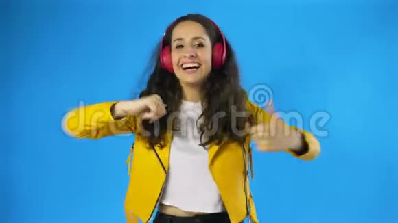 年轻女子穿着黄色夹克一边在蓝底工作室听耳机里的音乐一边跳舞视频的预览图