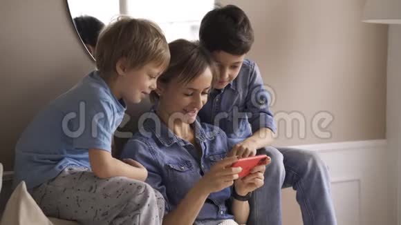 年轻的高加索女人有两个孩子玩智能手机在沙发上放松快乐的男孩喜欢在家里度过时光视频的预览图
