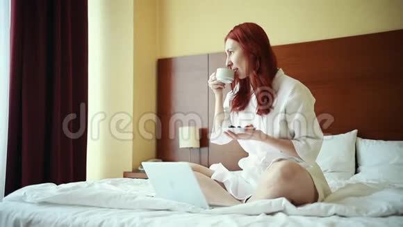 一个生姜女人坐在酒店房间的床上喝茶用笔记本电脑工作视频的预览图
