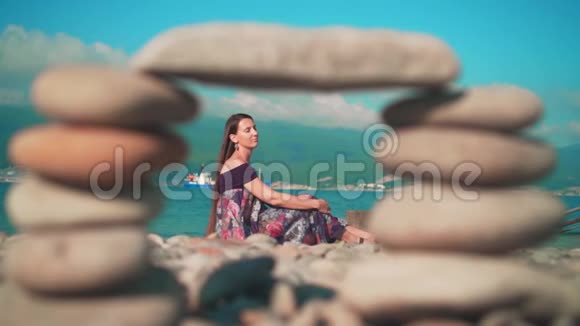 美丽的女孩穿着裙子坐在海滩上从一帧鹅卵石中拍摄的照片背后是大海群山视频的预览图