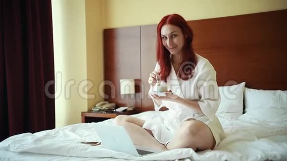 一个姜黄色的微笑女人坐在酒店房间的床上喝着茶视频的预览图