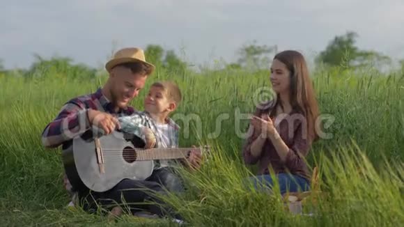 有魅力的小男孩正在学习弹吉他而爸爸妈妈则在家庭野餐放松时欢笑和欣赏视频的预览图