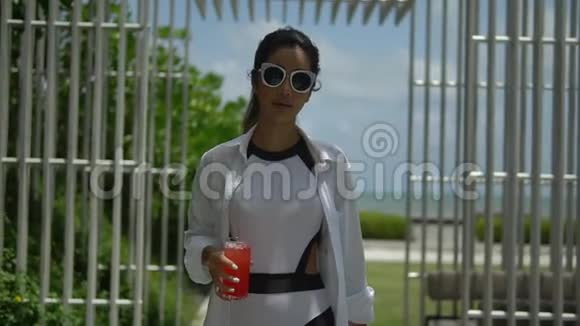 幸福的年轻女士穿着白色衬衫在热带酒店的阳光露台上散步手里拿着一杯红色的提神剂享受着视频的预览图
