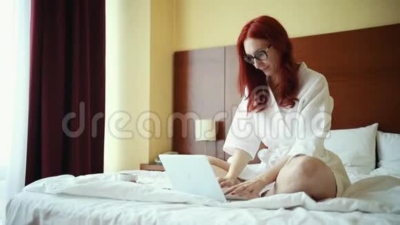 一个姜黄色的微笑女人坐在酒店房间的床上拿着笔记本电脑工作视频的预览图