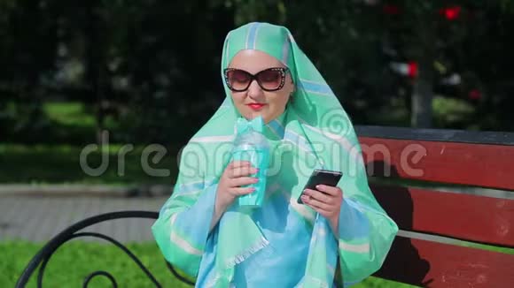 一位戴着浅色围巾和太阳镜的年轻穆斯林妇女坐在公园的长凳上喝着咖啡说话视频的预览图