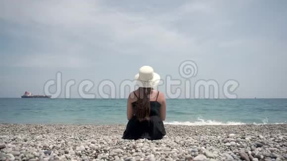 一个女孩穿着一件浅色连衣裙和一顶白色帽子坐在海滩上欣赏美丽的风景视频的预览图