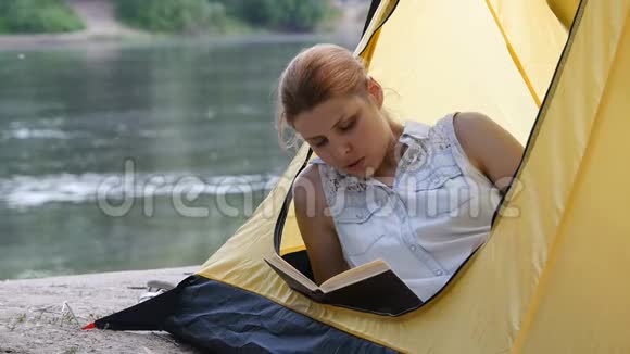 远足女孩在野营帐篷里看书人们生活在健康活跃的生活方式中背景中的河流和森林视频的预览图