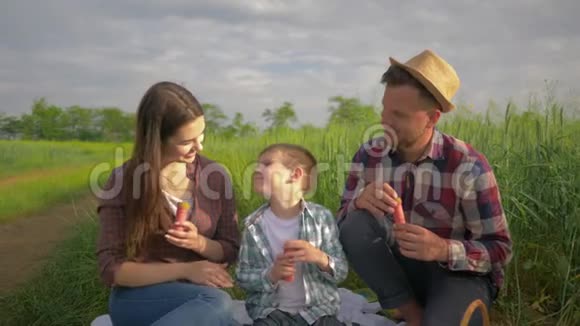 快乐的童年有趣的父亲和妈妈和男孩一起吹泡泡在绿色的田野里露天微笑视频的预览图