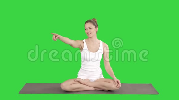 年轻漂亮的女人坐在莲花的姿势上指着什么或某人在绿色的屏幕上说话视频的预览图