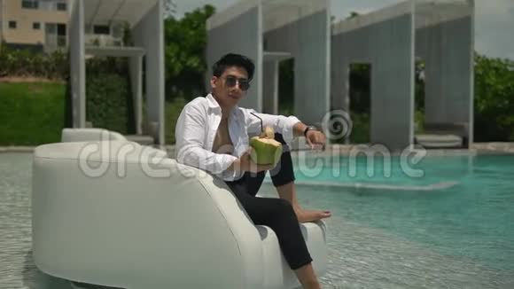 阳光明媚的一天穿着白色衬衫和黑色裤子的快乐年轻人在泳池边休息手里拿着椰子和稻草视频的预览图