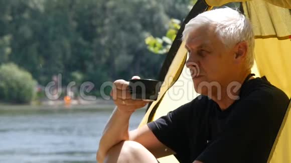 年长的灰发露营者坐在帐篷里喝茶快关门河流和森林的背景徒步旅行视频的预览图