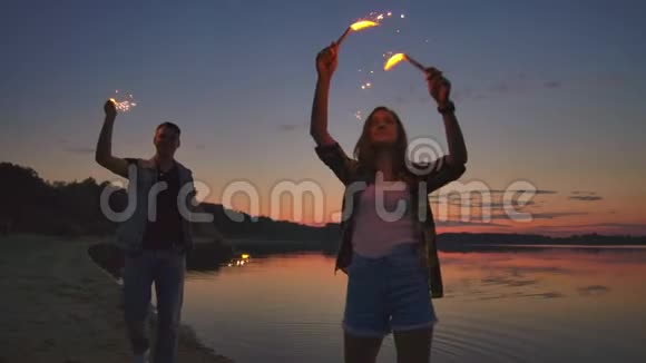 年轻快乐的多民族夫妇手持燃烧着闪闪发光的蜡烛日落时在海边奔跑慢镜头视频的预览图