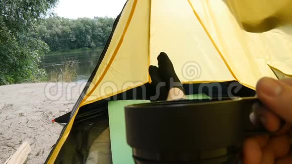徒步旅行者坐在露营帐篷里喝茶或咖啡独自旅行的独奏游客把腿伸直视频的预览图